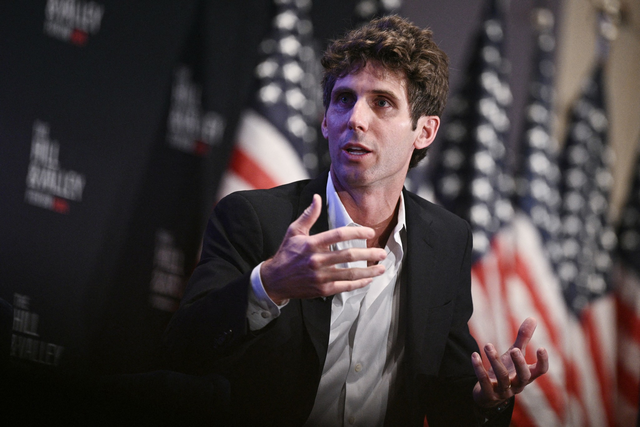 A man with curly hair in a dark suit gestures while speaking, with multiple blurred American flags in the background.